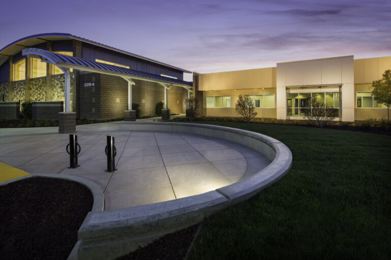 Modern building entrance with curved canopy, lit windows, and landscaped courtyard at dusk