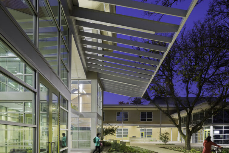 Modern glass building courtyard at dusk with a canopy, trees, and people walking and biking
