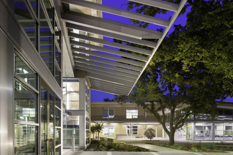 Modern glass building courtyard at dusk with a large tree and overhead white canopy beams