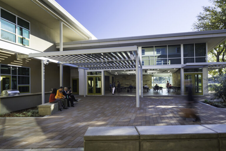 Modern building courtyard with people sitting under a pergola and others in a glass-walled patio