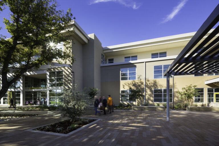 Modern campus building courtyard with trees, a shaded walkway, and a few people walking under a blue sky