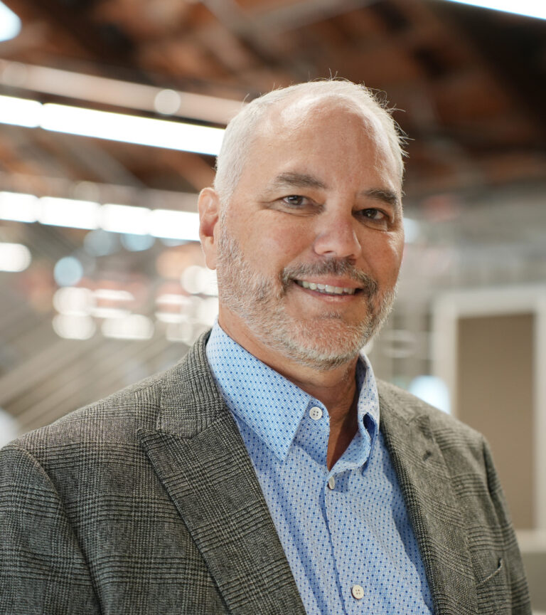 Smiling middle-aged man with gray beard in a blazer and blue shirt, standing indoors with a blurred background