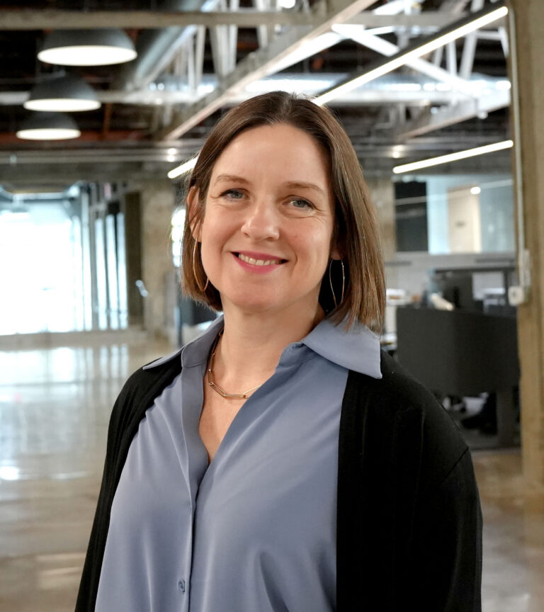 Smiling woman in a modern office with industrial lighting, wearing a blue blouse and black cardigan